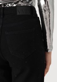 Črne jeans hlače iz denim materiala z teksturirano površino, manjšimi zakovicami, eno zadnjo žepom in črnim usnjenim oznako VMD na pasu.