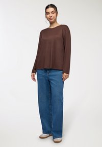 Brązowa bluzka z długim rękawem o luźnym kroju, w połączeniu z szerokimi niebieskimi dżinsami i jasnymi butami. Gładka tekstura materiału.