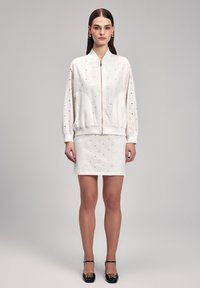 Set composto da bomber bianco e gonna con design a occhiello, dotato di zip e polsini a coste. Il modello indossa scarpe nere con un accento dorato.