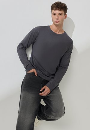 Maglietta grigia a maniche lunghe in morbido tessuto, con scollatura a girocollo, abbinata a jeans in denim scuro e sneakers bianche, creando un look casual.