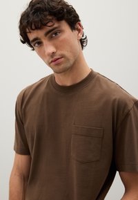 T-shirt in cotone marrone con maniche corte e scollatura rotonda. Presenta una tasca sul petto e una texture liscia e morbida. Design semplice.