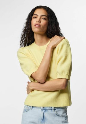 Mujer con cabello largo y rizado, vistiendo un suéter amarillo de manga corta y vaqueros azul claro, posando con un brazo cruzado sobre su cuerpo.