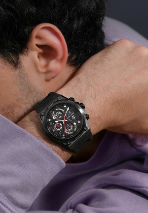 Black sport watch with a textured bezel, multi-layered dark dial, red accents, and a rubber strap, worn on a wrist.