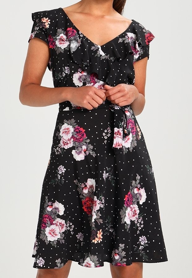 Femme portant une robe noire midi avec des pois blancs et des motifs floraux roses, rouges et blancs, dotée d'un col froncé et d'une ceinture à la taille.