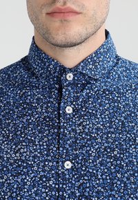 Camicia button-up in tessuto blu scuro con un piccolo motivo floreale blu. Presenta un colletto a punta e bottoni bianchi lungo la parte anteriore.