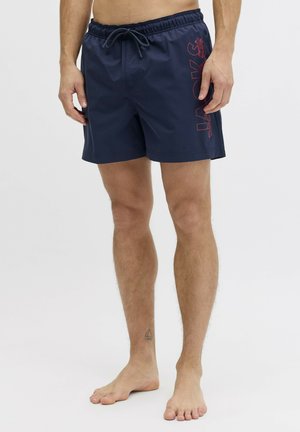 Homme portant un short de bain bleu marine avec le texte rouge "JACK & JONES" sur le côté, debout pieds nus sur un fond blanc.