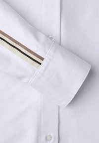 Biały mankiet koszuli wykonany z gładkiej tkaniny, z brązowym, kremowym i czarnym paskowanym akcentem wzdłuż rękawa. Zawiera guzik jako detal.