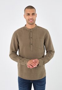 Braunes, knöpfbares, langärmliges Hemd mit rundem Kragen, einer Brusttasche und strukturiertem Stoff. Kombiniert mit blauen Jeans. Einfarbiger Hintergrund.
