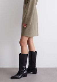 Bottes noires à talons hauts avec une finition brillante, design à bout effilé et une silhouette à hauteur de genou. Associées à une robe texturée vert clair.