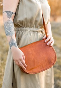 Hnědá kožená křížová taška zaobleného tvaru, hladkého povrchu a prošitými okraji, držena rukou blízko béžových šatů.