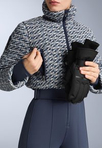 Mornarice i bijela pletena jakna s visokim ovratnikom, koja ima džep sa zatvaračem. Crne rukavice u jednoj ruci. Uske mornarice hlače.