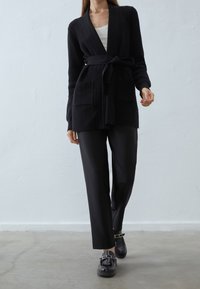 Schwarze gerippte Strickjacke mit einem Bindegürtel, ausgestattet mit zwei Vordertaschen. Kombiniert mit schwarzen Hosen und klobigen schwarzen Schuhen. Glatte Textur.