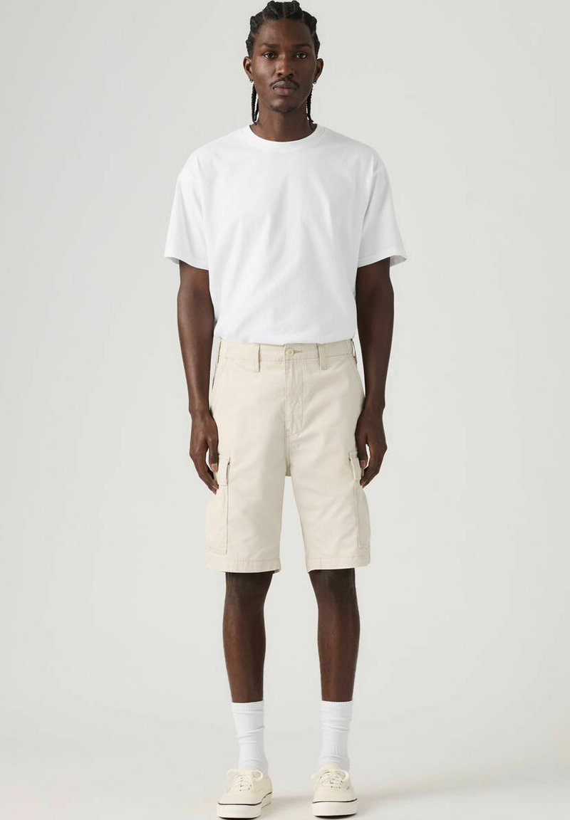 T-shirt en coton blanc, short cargo beige avec poches latérales, chaussettes jusqu'à la cheville et baskets légères, debout contre un fond uni.