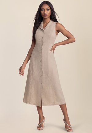 Woman with long dark hair wearing a sleeveless beige button-front midi dress and silver high-heeled sandals, standing with hand on hip.