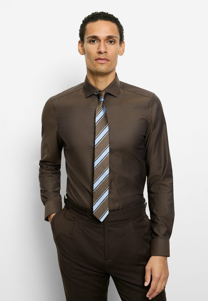 Braun gemustertes Hemd mit Haifischkragen, kombiniert mit einer gestreiften Krawatte in Blau- und Brauntönen sowie dunkelbraunen Anzughosen.