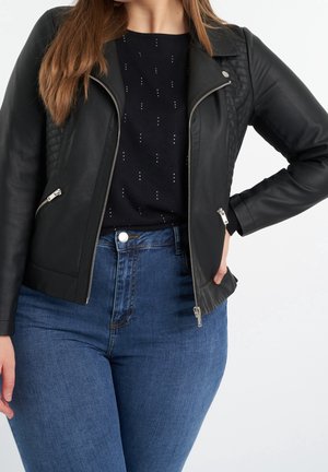 Femme portant une veste en cuir noire zippée sur un top noir à motifs, associée à un jean bleu taille haute, main reposant sur la hanche.