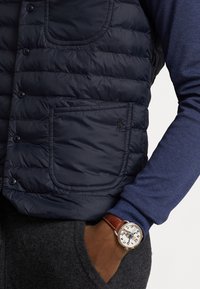 Granatowa pikowana kamizelka z przeszywanym wzorem, przednią kieszenią i zapięciem na guziki, noszona na niebieskiej koszuli z długim rękawem. Na nadgarstku widoczny jest zegarek.