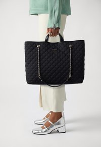 Personne tenant un sac cabas matelassé noir de la marque Guess, portant un pantalon blanc, une veste vert menthe, et des chaussures Mary Jane à talon carré argentées.