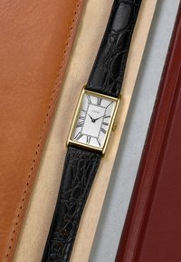 Rechteckige goldene Timex-Uhr mit weißem Zifferblatt, schwarzen römischen Ziffern und schwarzem strukturiertem Lederarmband zwischen braunen und roten Lederflächen.