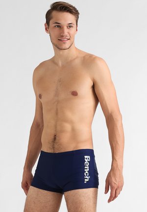 Marine Badehosen aus glattem, dehnbarem Material, mit einem weißen "Bench"-Logo, das vertikal auf der linken Seite aufgedruckt ist.