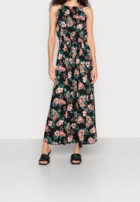 Frau in einem schwarzen Maxikleid mit floralem Halsausschnitt, verziert mit pinken und grünen Blumen, kombiniert mit schwarzen, geflochtenen Sandalen mit offenem Zeh und Absatz.