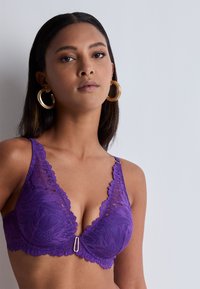 Soutien-gorge en dentelle violette avec motif floral, décolleté plongeant, bords festonnés et détail en métal doré au centre. Texture lisse.