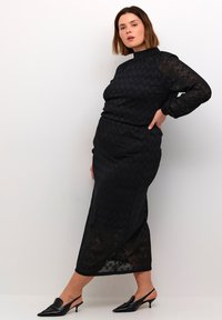 Svart spetsklänning med hög halsringning, långa ärmar och en åtsittande silhuett. Har ett subtilt mönster och paras med svarta slingback-klackar.
