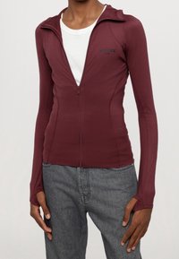 Burgundská zipová bunda s dlouhými rukávy, přiléhavý střih a otvory pro palce. Hladká textura s logem na levém prsu.