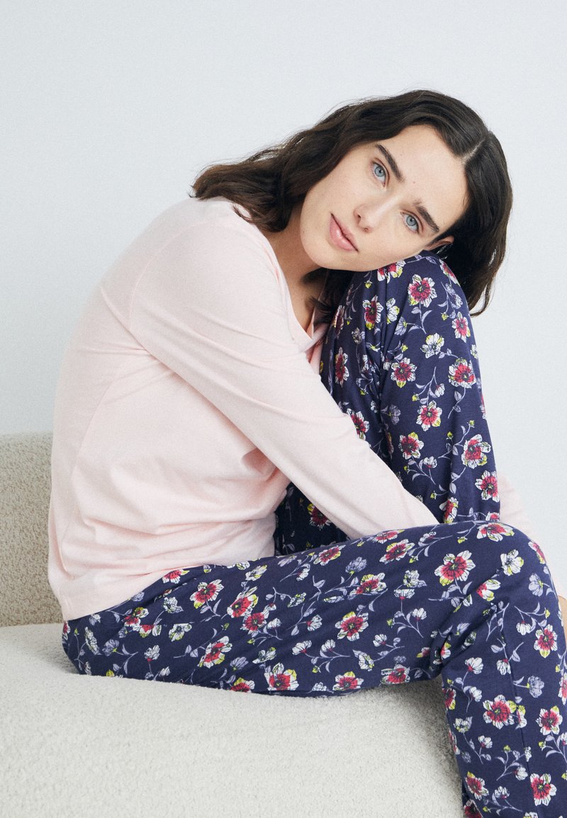 Ljusrosa långärmad topp i kombination med marinblå pyjamasbyxor med blommönster som har röda och vita blommor. H sitting pose på en ljus yta.