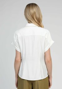 Femme blonde se tenant de dos, portant un chemisier blanc à manches courtes et un pantalon vert olive, devant un fond clair et uni.