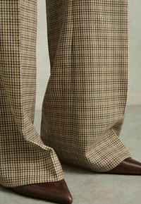 Breda byxor i ett brun houndstooth-mönster, med en texturerad tyg och spetsiga bruna klackar, som visar upp rena linjer och skräddarsydd finish.
