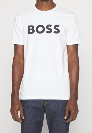Person trägt ein weißes kurzärmeliges T-Shirt mit „BOSS“ in großen schwarzen Buchstaben auf der Brust, kombiniert mit dunkelblauen Jeans.