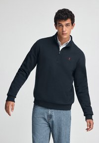 Sudadera negra con cremallera y cuello alto, puños acanalados y cintura de canalé. Presenta un pequeño logo rojo en el pecho. Llevado sobre una camisa con cuello.