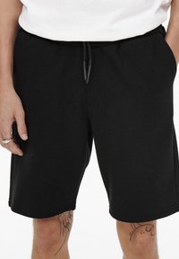 Pantaloni shorts neri in cotone con vita elasticizzata, dettaglio con coulisse e tasche laterali; texture liscia e design lungo fino al ginocchio.