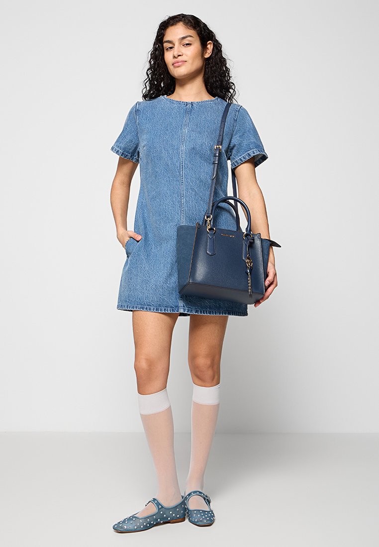 Abito in denim con maniche corte, colore azzurro chiaro, zipper frontale e due tasche laterali. Porta una borsa a mano blu scuro con un portachiavi.