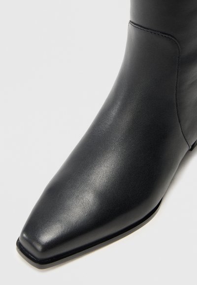 Čierna kožená členková čižma s tvarovanou špičkou, hladkým povrchom a minimálnym prešívaním. Podrážka má mierne zakrivenie pre ergonomické prispôsobenie.