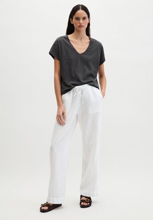 Frau stehend, trägt dunkelgraues V-Ausschnitt-T-Shirt mit kurzen Ärmeln, weiße weit geschnittene Hose mit Kordelzug und braune Sandalen mit goldener Schnalle.