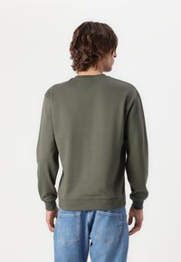 Felpa verde oliva con maniche lunghe, scollo rotondo e polsini a costine. Indossata con jeans azzurri chiari caratterizzati da un dettaglio di cucitura sottile sul retro.