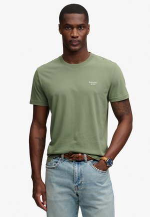 Mężczyzna w oliwkowej koszulce Superdry, jasnoniebieskich jeansach, brązowym plecionym pasku i zegarku na nadgarstku, stoi z jedną ręką w kieszeni.