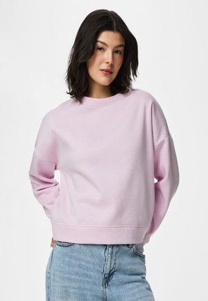 Giovane donna con capelli scuri fino alle spalle che indossa una felpa rosa chiaro e jeans blu, in piedi con le mani dietro la schiena.