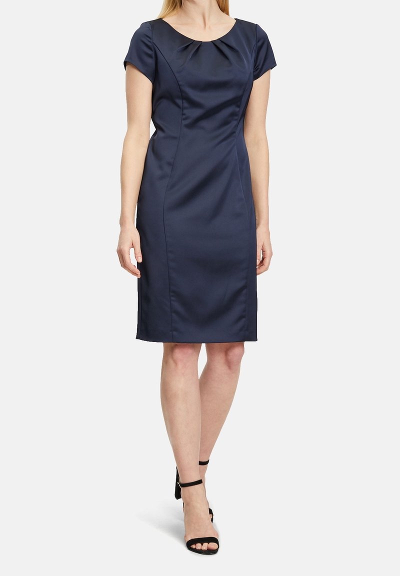 Femme portant une robe bleu marine ajustée au genou avec des manches courtes et des talons noirs à bride à la cheville, debout devant un fond uni.