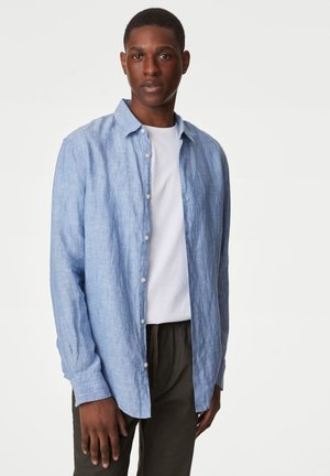 Camisa de botones de manga larga en azul claro con textura suave, cuello y botones blancos, llevada sobre una camiseta blanca y pantalones oscuros.