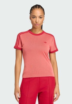 Ung kvinde med fletninger iført en rød Adidas t-shirt og matchende røde bukser, stående mod en ensartet lys baggrund.