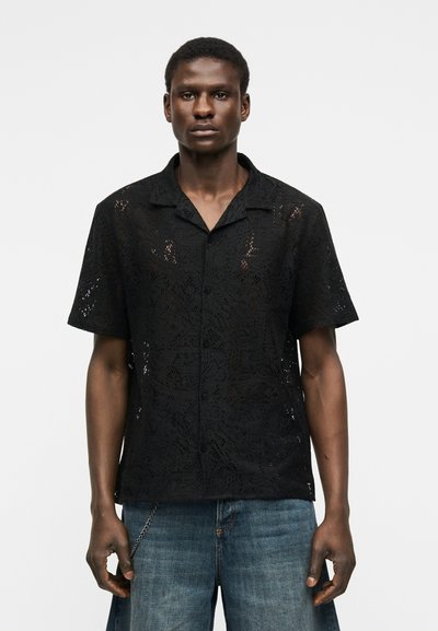 Άνδρας που φοράει μαύρο πουκάμισο με κουμπιά και κοντά μανίκια από δαντέλα και φαρδιά μπλε τζιν, με φόντο απλό άσπρο.