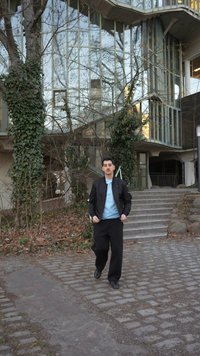 Uomo con giacca nera e camicia azzurra che cammina su un sentiero di ciottoli vicino a scale e un edificio di vetro coperto di edera.