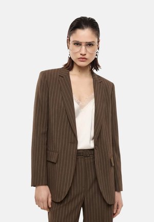 Donna che indossa un completo marrone a righe con pantaloni coordinati, camicetta bianca con rifiniture in pizzo, grandi occhiali trasparenti e orecchini vistosi, su sfondo neutro.