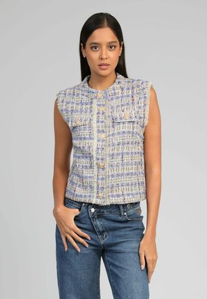 Jeune femme aux longs cheveux foncés portant un gilet texturé sans manches boutonné et un jean bleu, posant devant un fond gris uni.