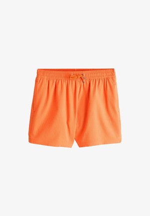 Bărbat fără tricou, cu păr creț, purtând ochelari de soare negri și șorturi de baie portocalii strălucitori, stând în fața unui perete bej texturat.