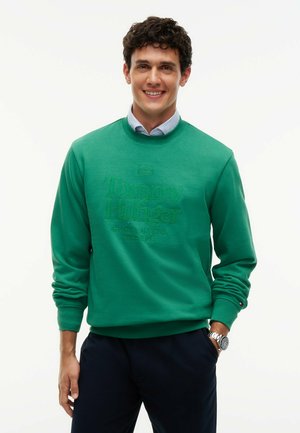 Uomo con capelli ricci che indossa una felpa verde Tommy Hilfiger sopra una camicia a righe, pantaloni scuri e un orologio da polso, sorridente verso la fotocamera.