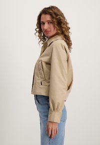 Beige cropped jack met een kraag, zijzak en knoopsluiting. Heeft lange mouwen en een gladde textuur. Gepaarde met blauwe jeans.